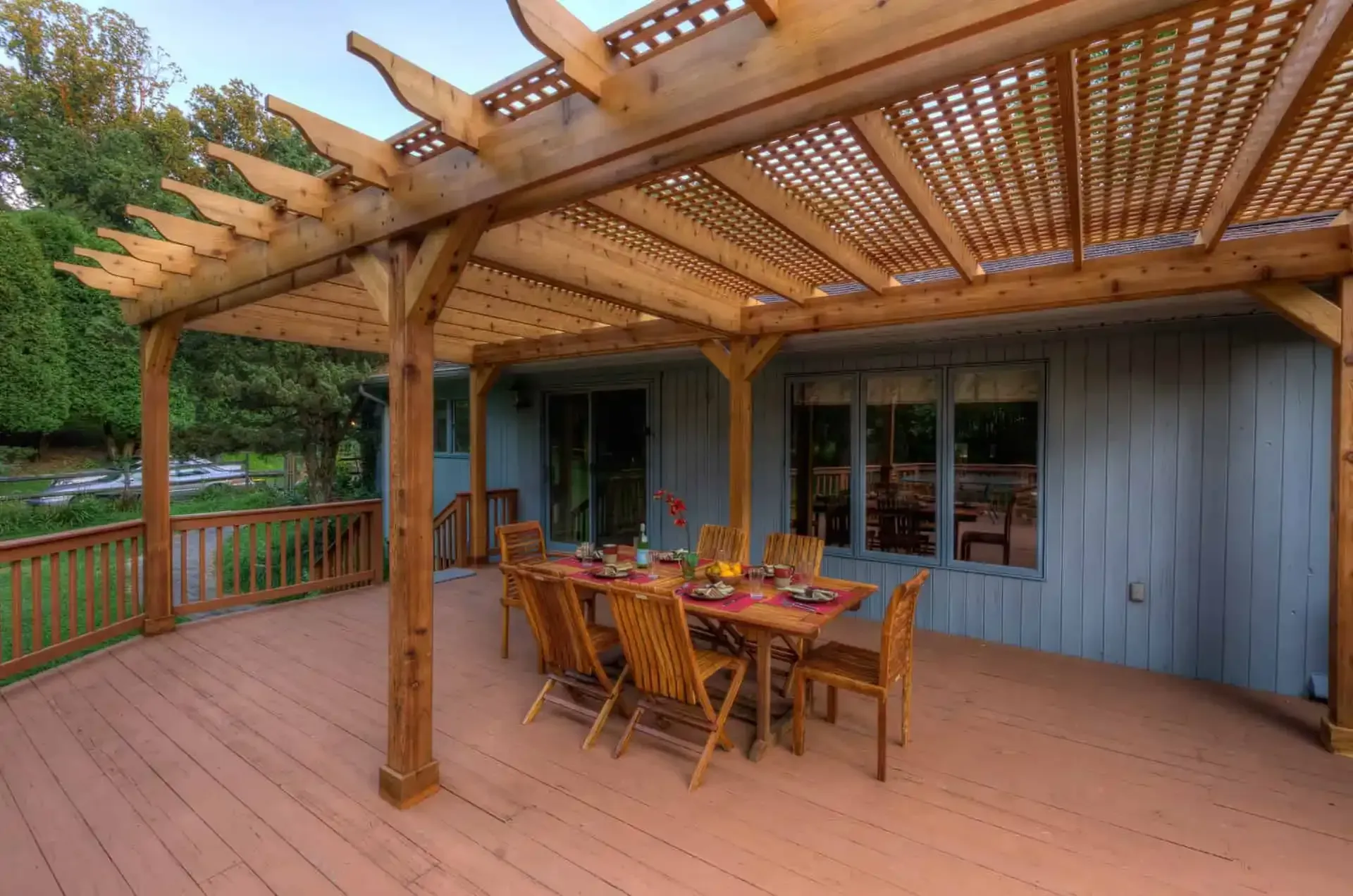 Wooden deck with pergola, dining table, and chairs beside a gray house in a wooded yard