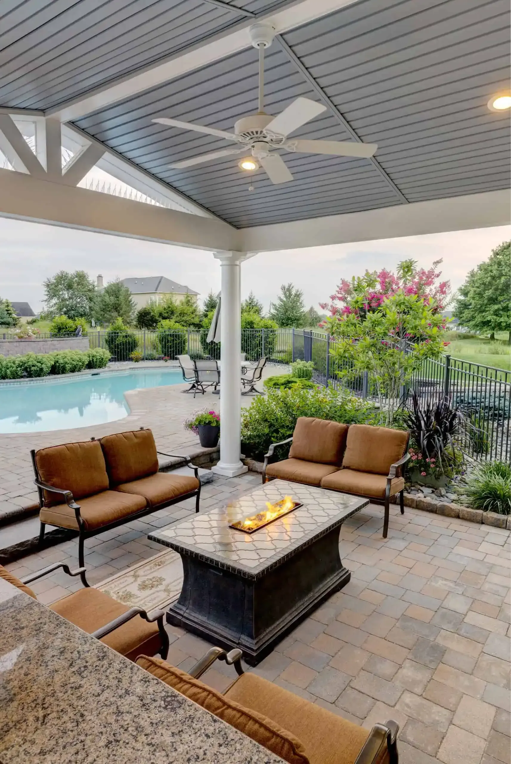 Covered patio with wicker seating around a fire table, pool, and lush garden view.