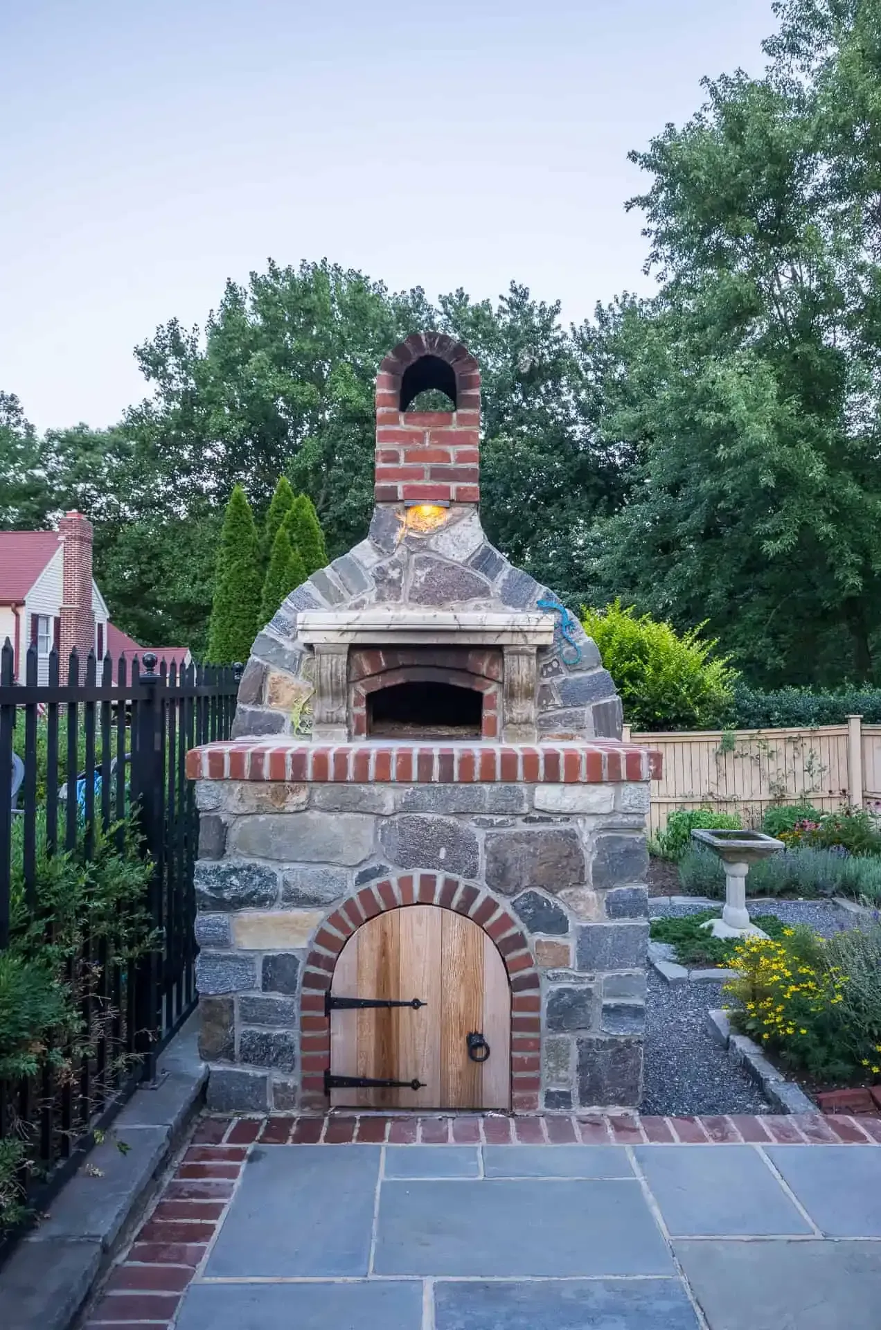 Outdoor brick and stone pizza oven with arched wooden door and chimney in a backyard patio