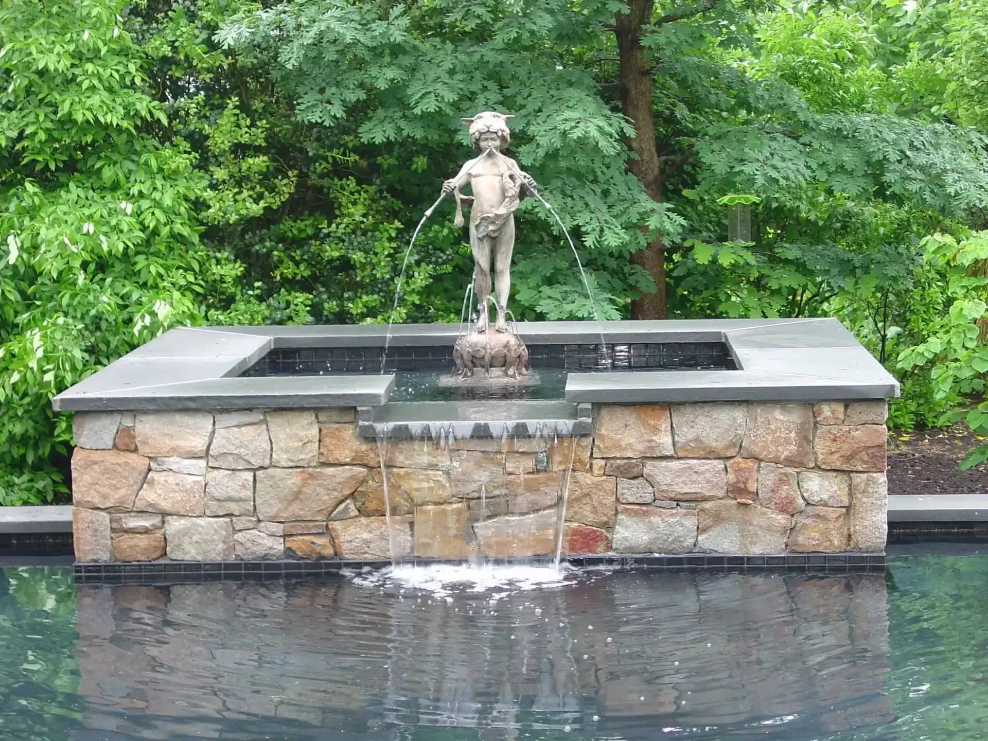 Small fountain with a standing statue atop a stone wall, set beside a reflecting pool and green trees.