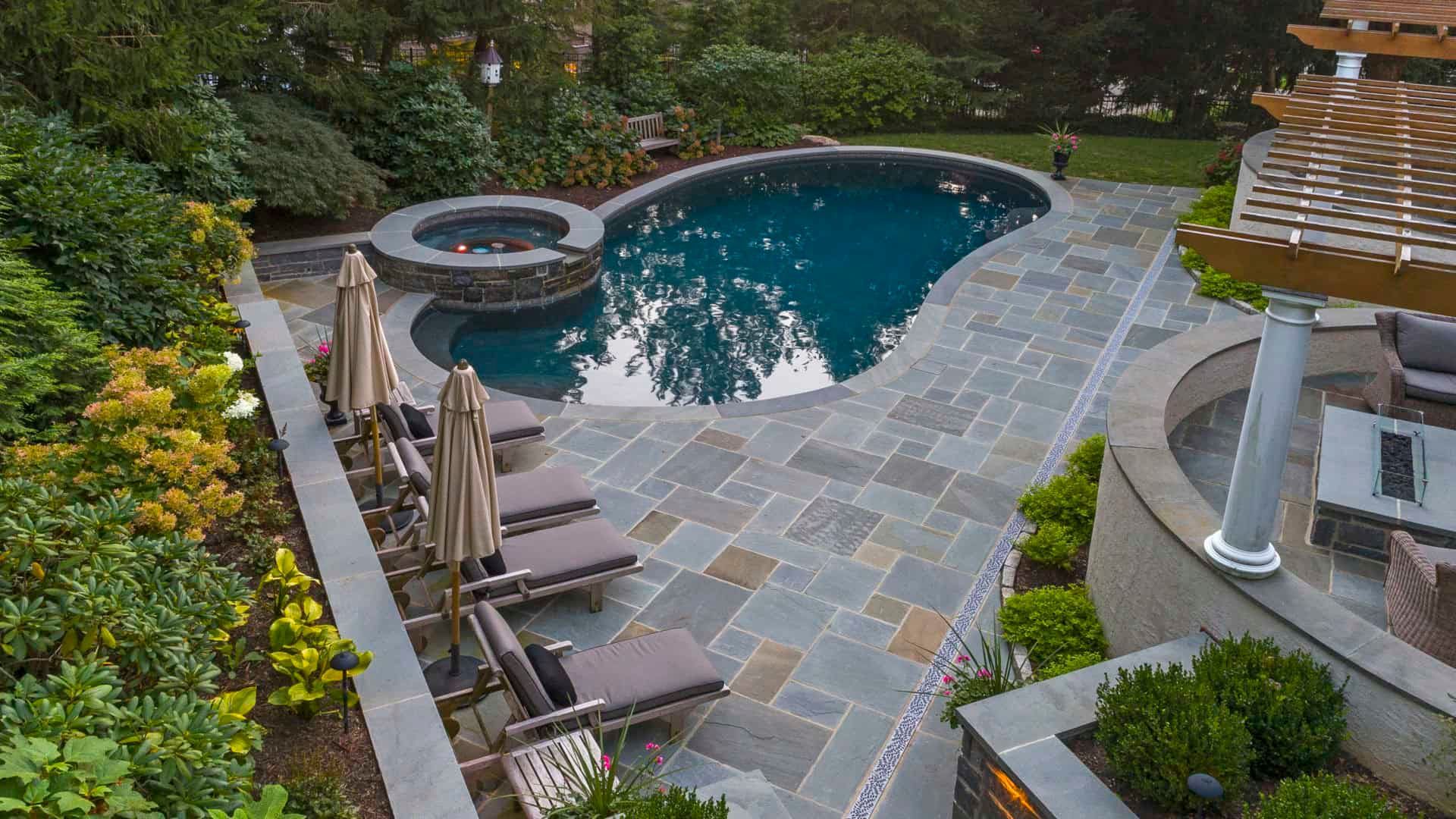 Backyard pool and hot tub with stone patio, lounge chairs, and surrounding greenery