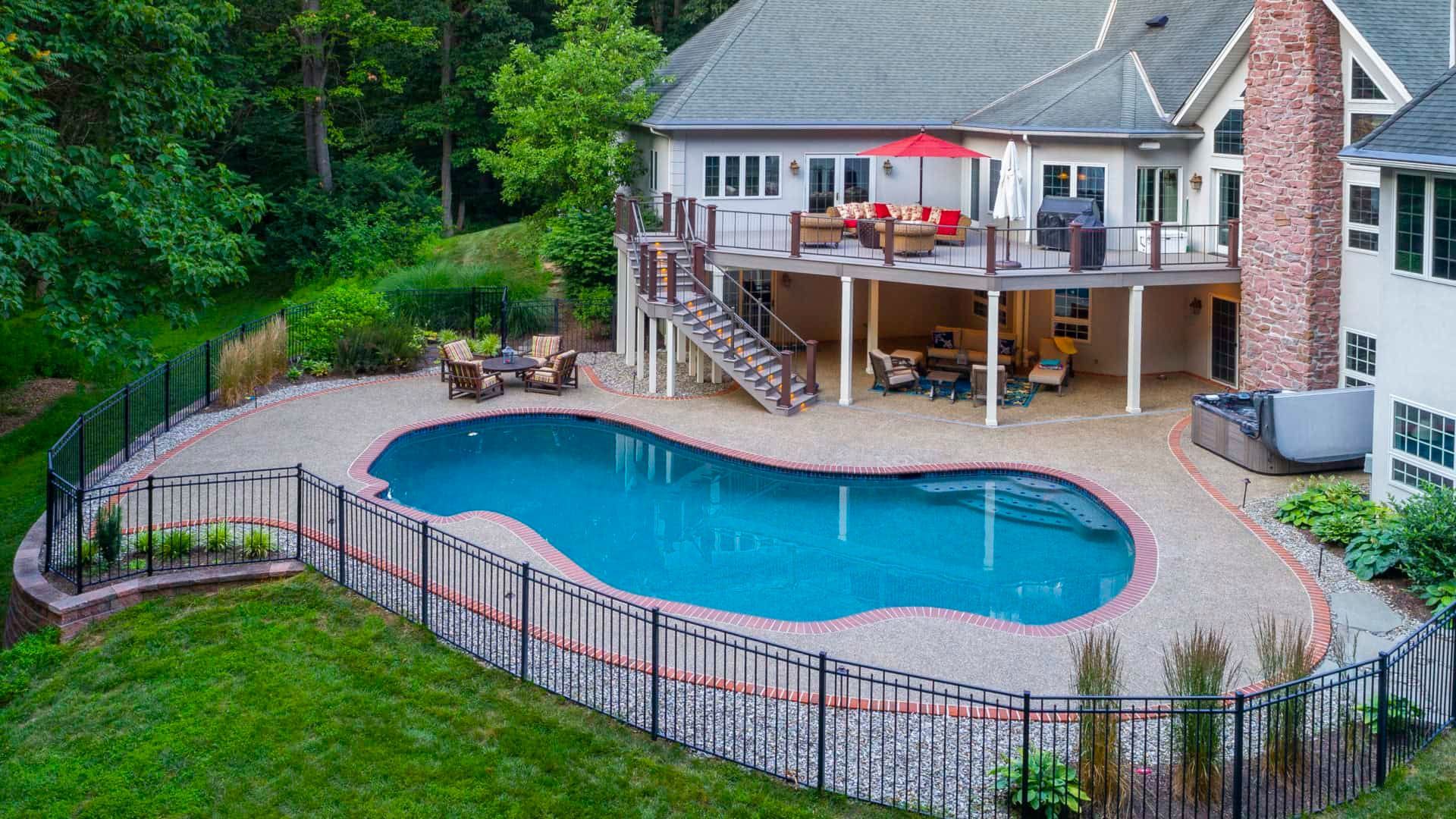 Backyard pool with a curved blue swimming pool beside a house and patio deck, bordered by a black fence