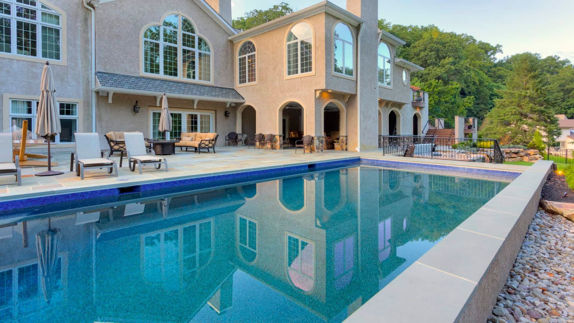 Luxury backyard with a large rectangular pool beside a stone patio and a two-story house.