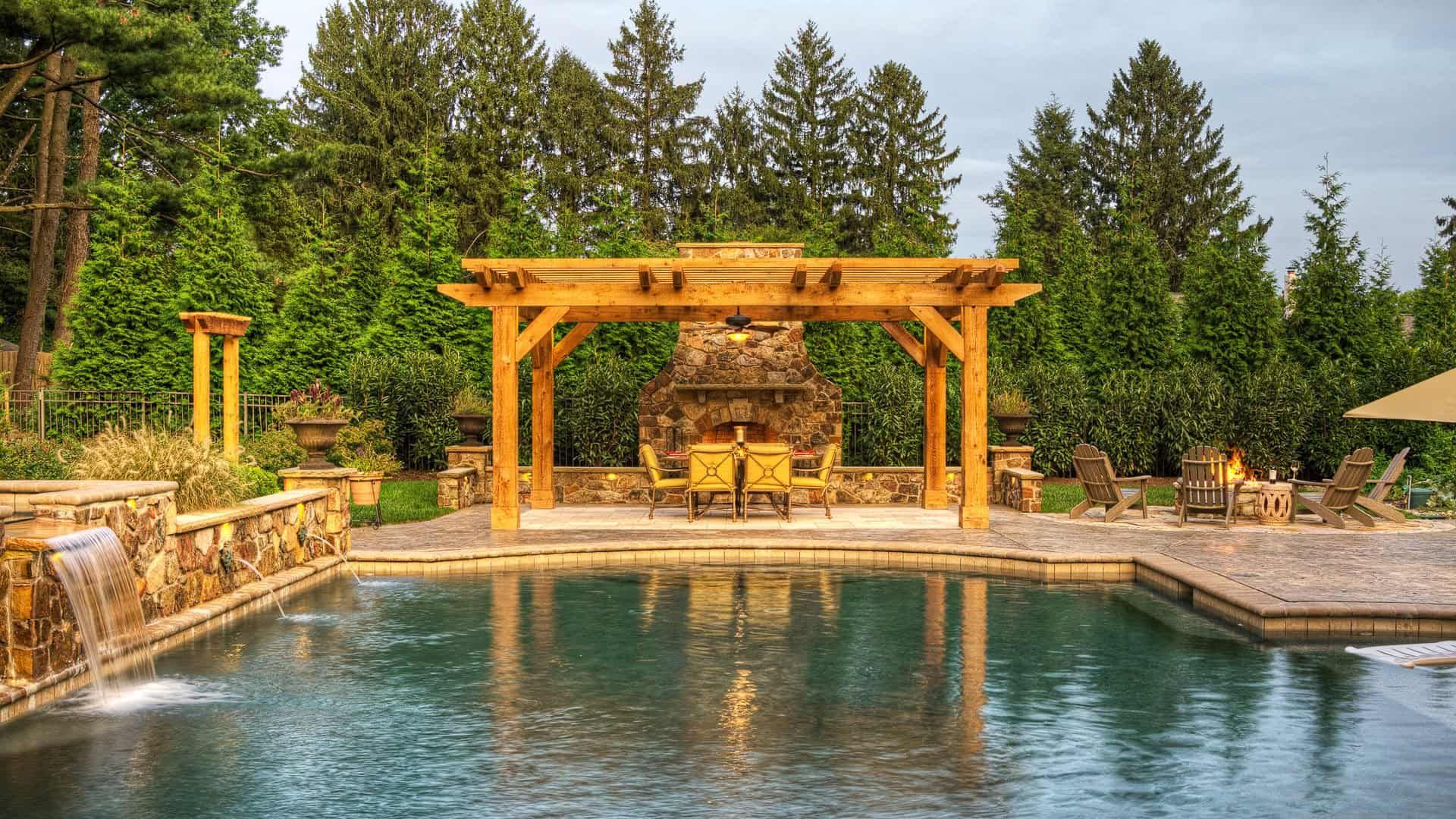 Backyard pool with wooden pergola, stone fireplace, and forest backdrop at sunset