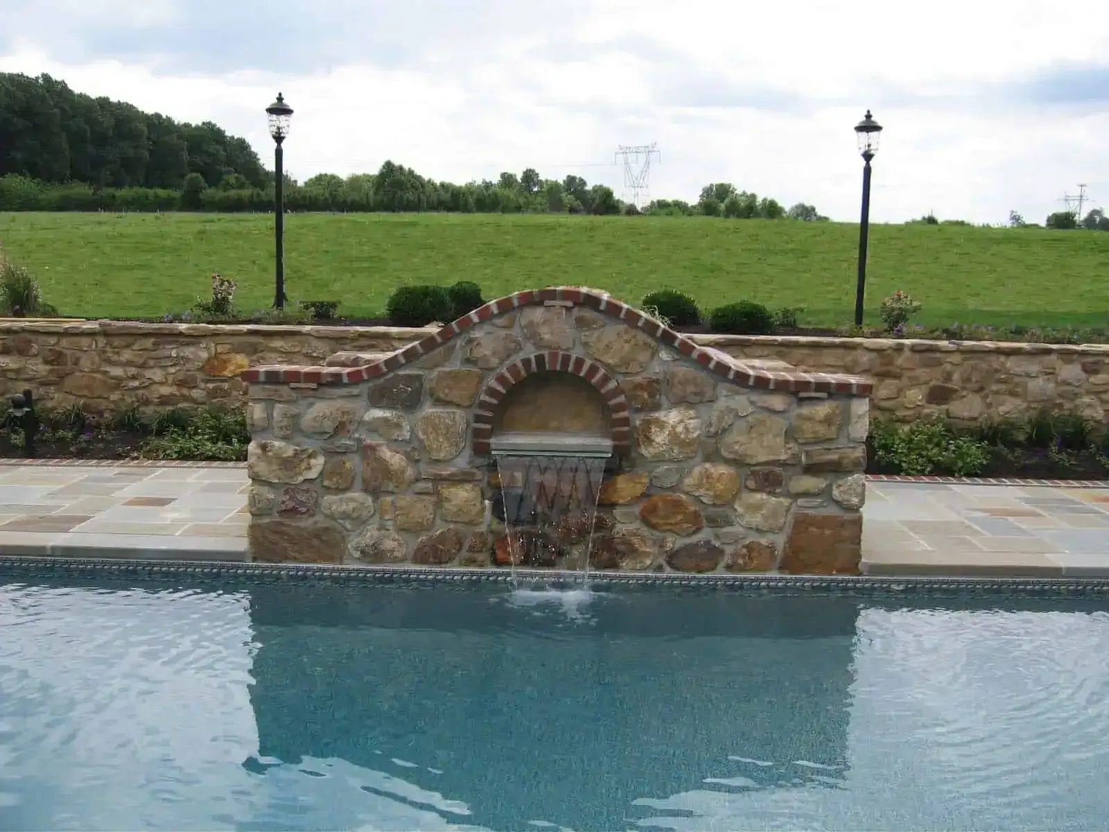 Stone fountain wall beside a blue pool, with green field and lampposts in the background.