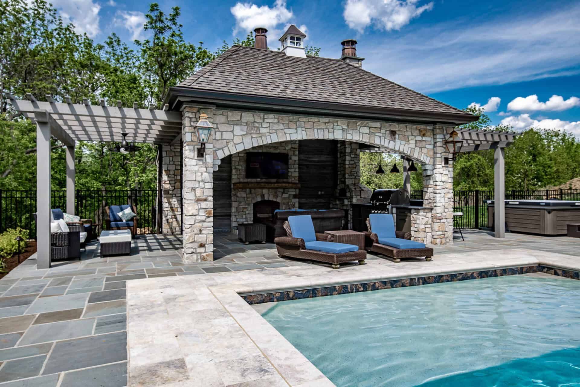 Stone poolside cabana with lounge chairs, pergola, and blue swimming pool in a landscaped backyard