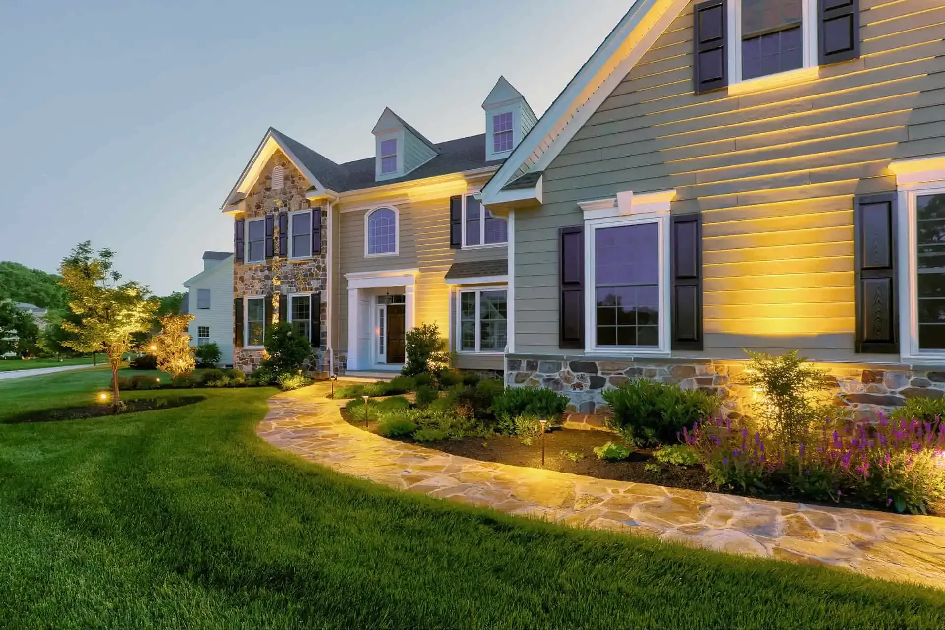 Elegant suburban house at dusk with warm exterior lights, manicured lawn, and landscaped garden path
