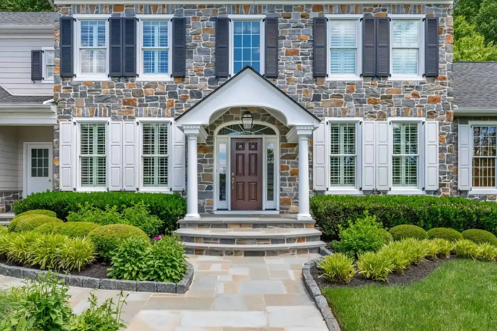 Stone house facade with black shutters, arched front door, and manicured hedges