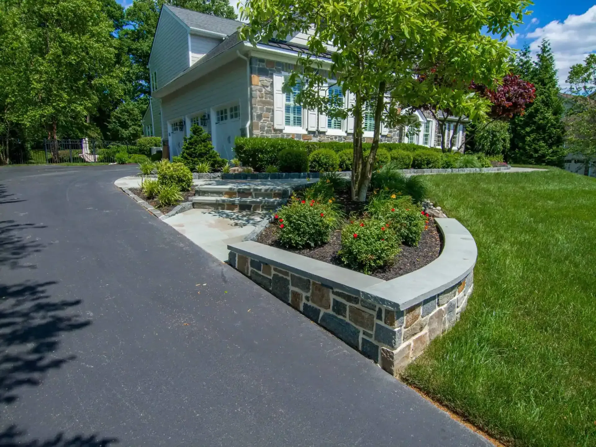 Curved stone planter beside a paved driveway and white house with green lawn and trees
