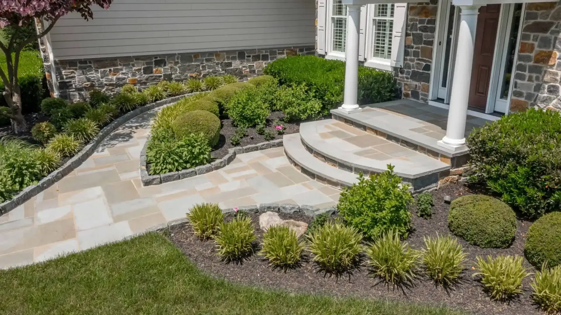Front yard landscaping with stone walkway, curved steps, and trimmed shrubs beside a house entrance
