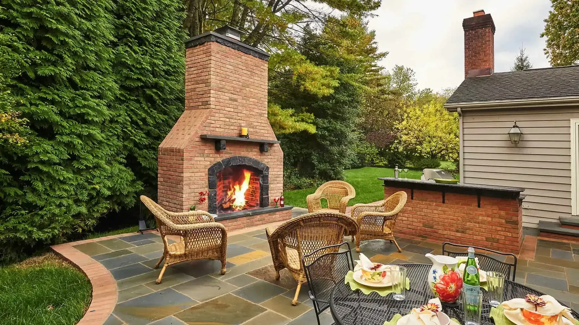 Brick outdoor fireplace beside wicker chairs and a table on a stone patio with garden greenery