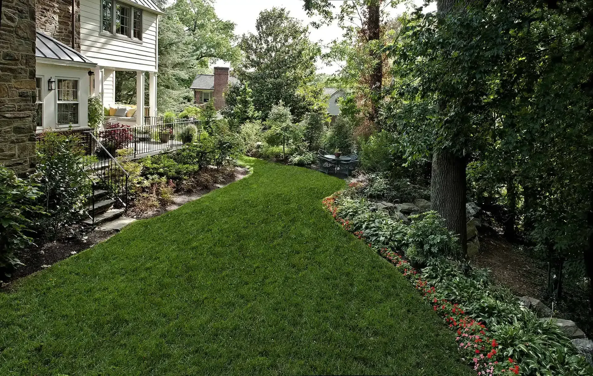 Lush green backyard garden path beside a house, lined with flowers and dense trees