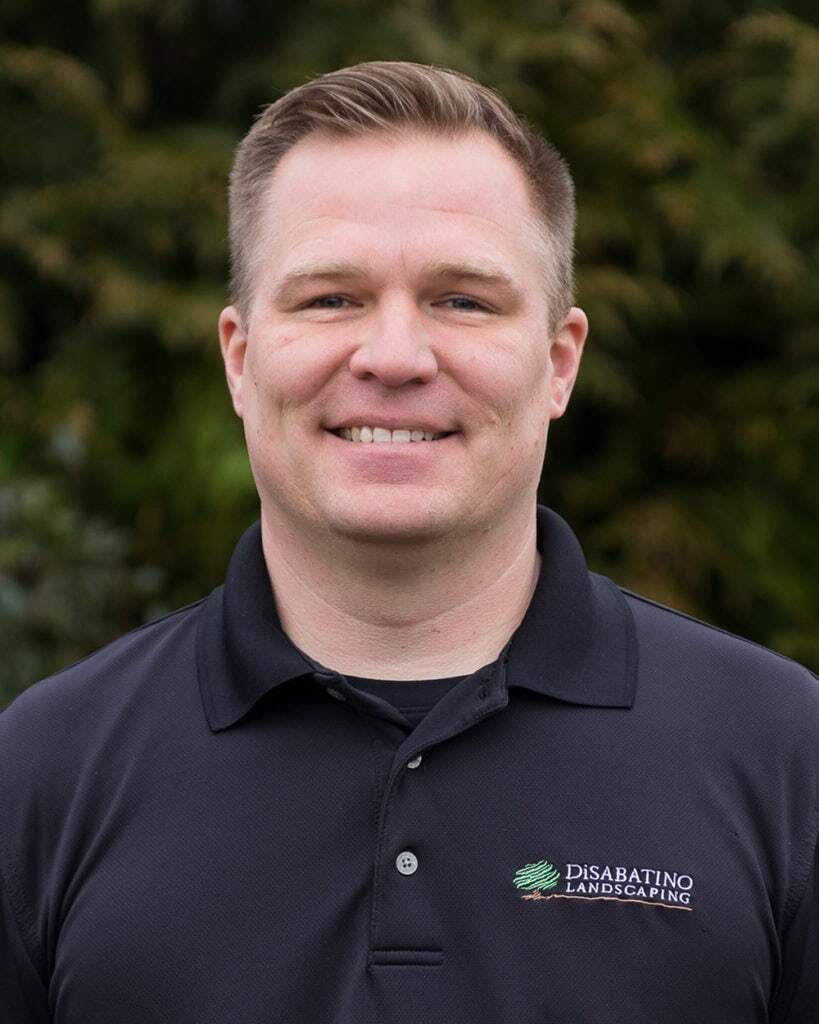 Smiling man in a dark navy polo shirt standing outdoors with blurred greenery behind him