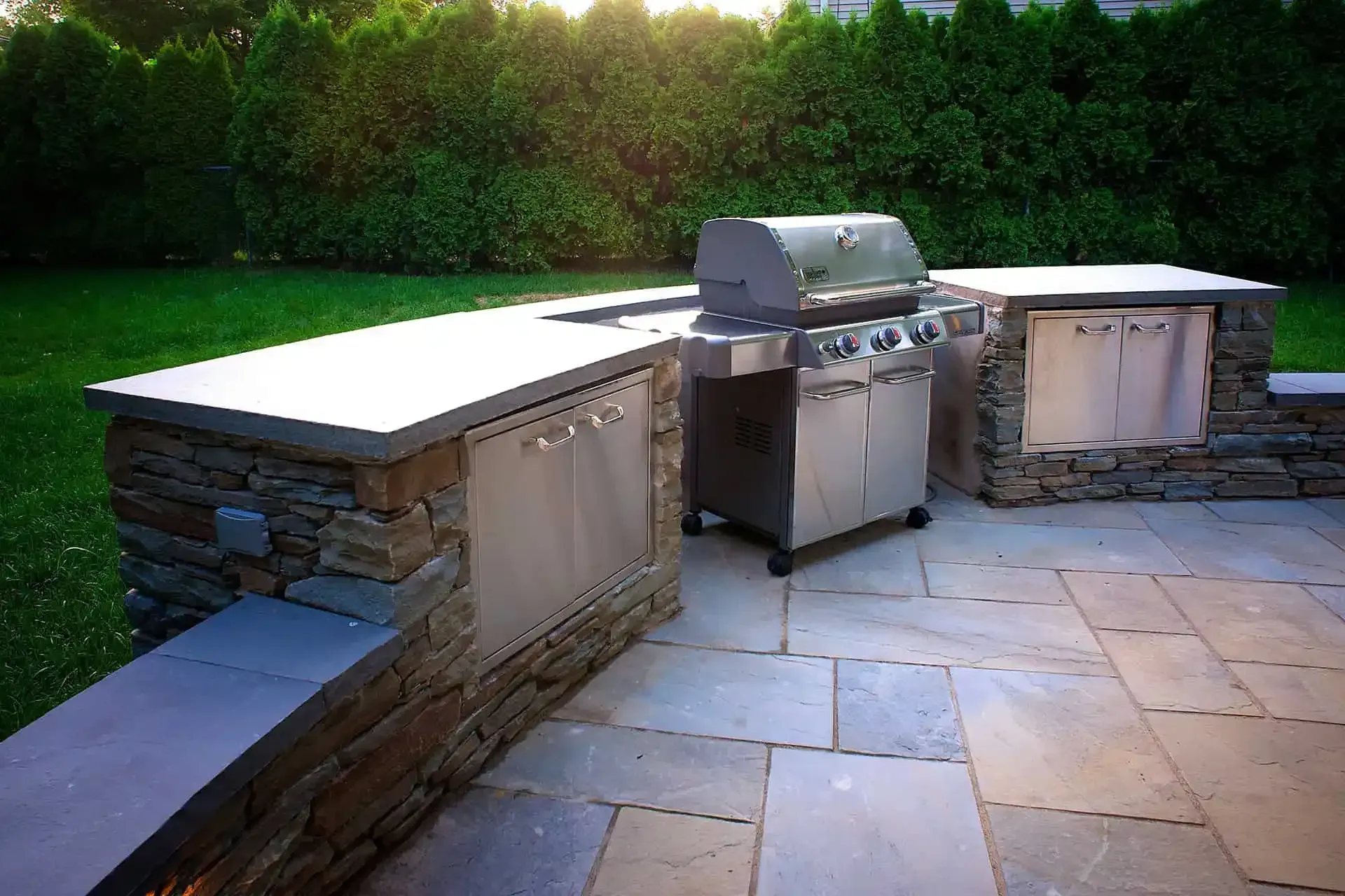 Outdoor stone patio with built-in grill and cabinets, bordered by green hedges