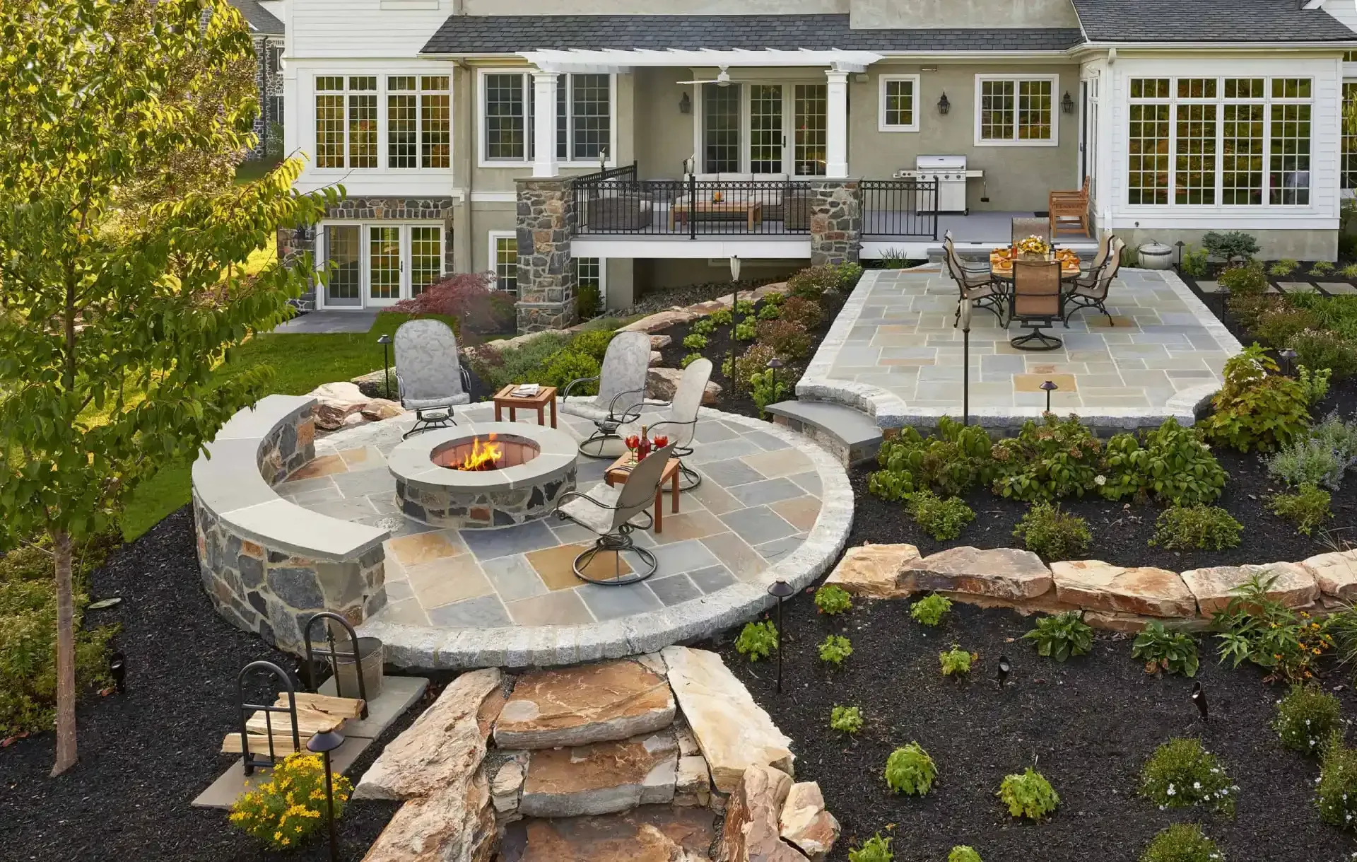 Patio with circular stone fire pit, chairs, and outdoor dining area in landscaped backyard behind a house