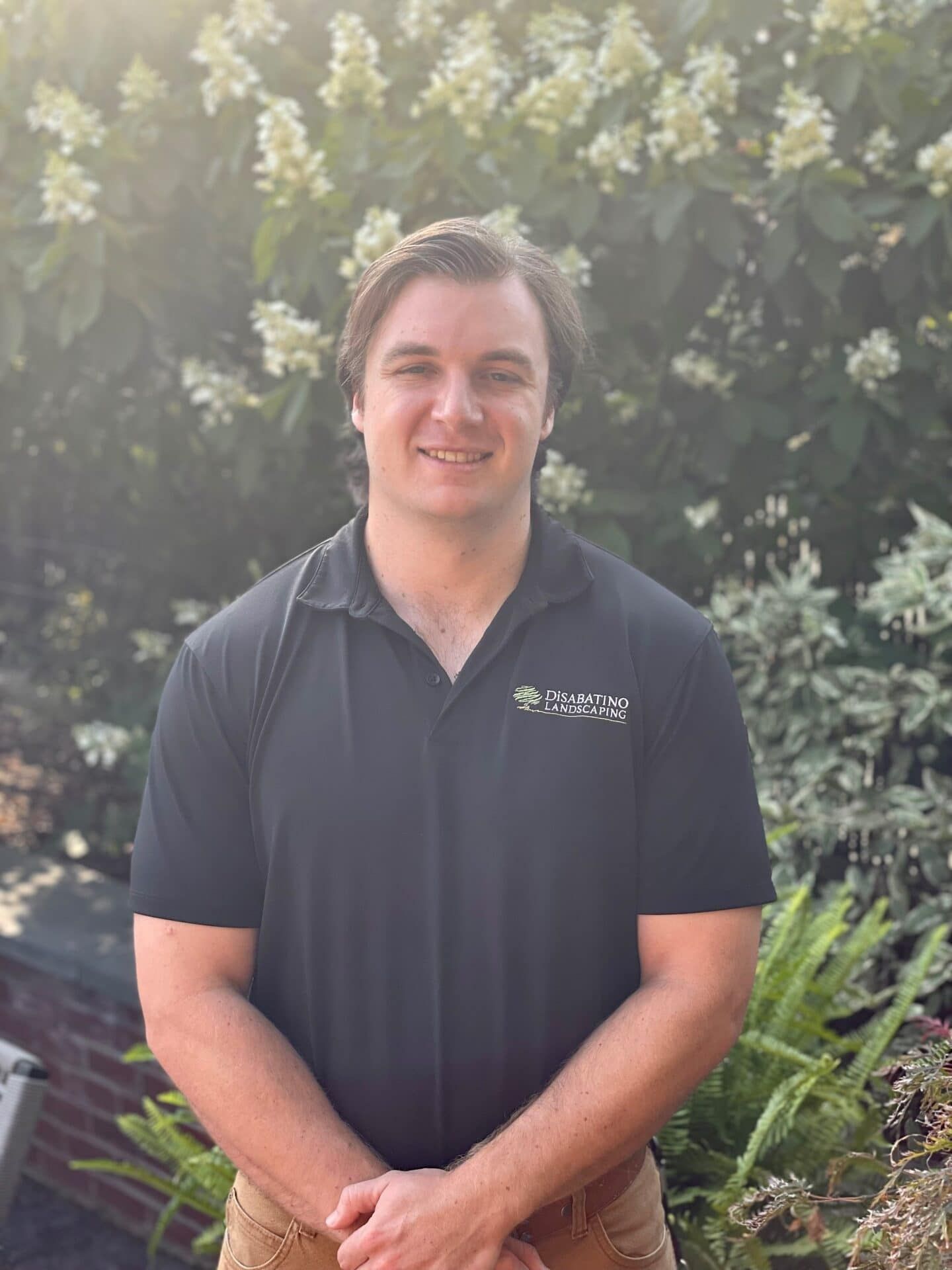Smiling person in a dark polo shirt standing outdoors in front of flowering bushes.