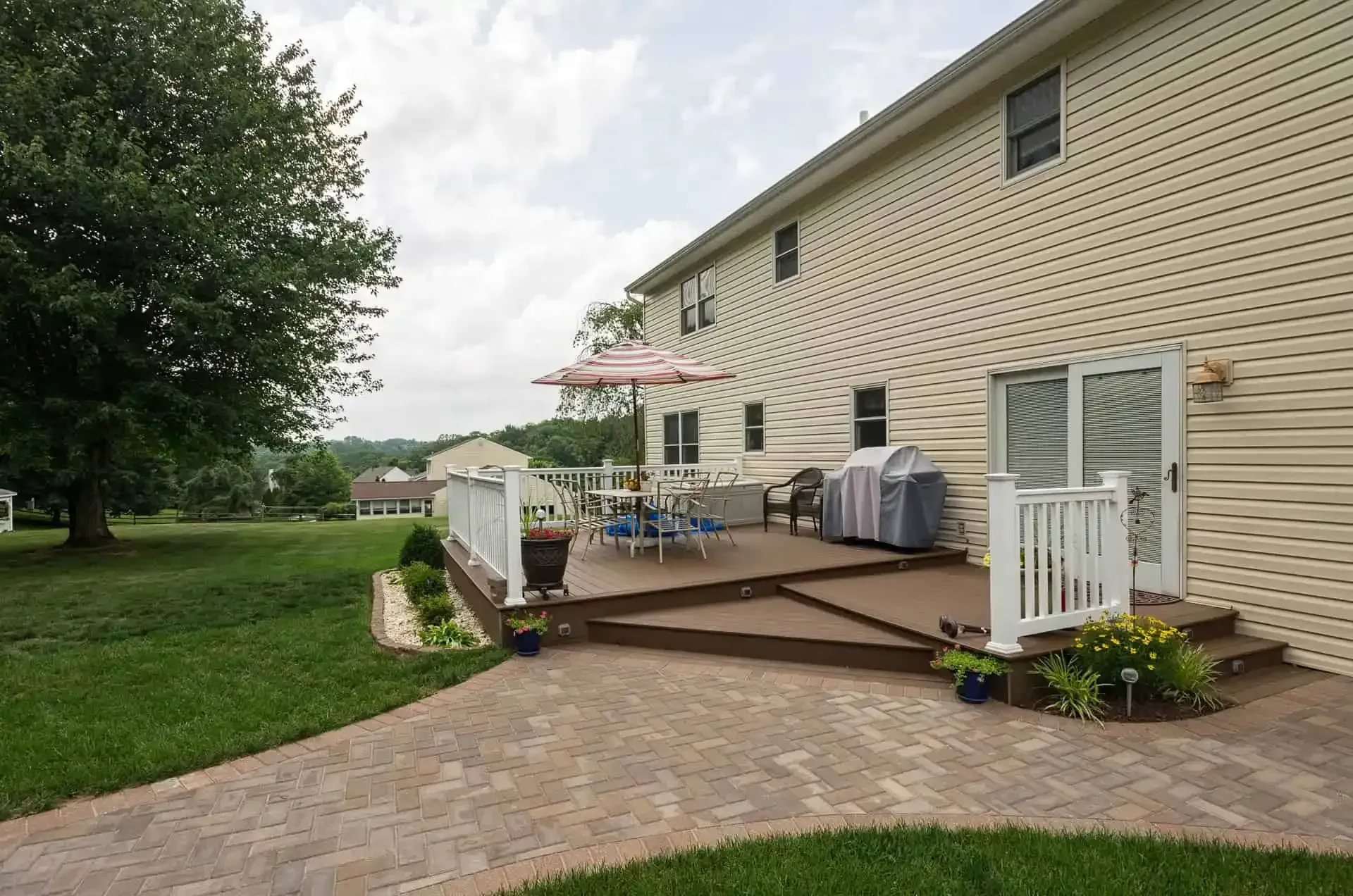Backyard deck beside beige house with patio furniture and grill, bordered by green lawn and a brick walkway