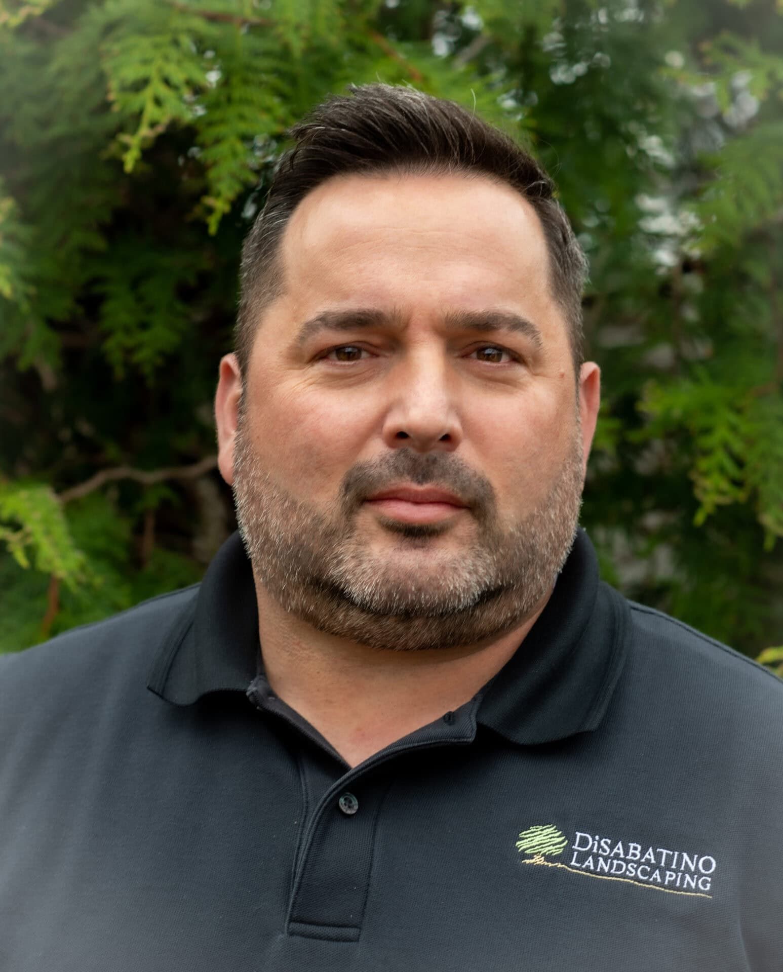 Portrait of a man in a black polo shirt standing outdoors against green trees
