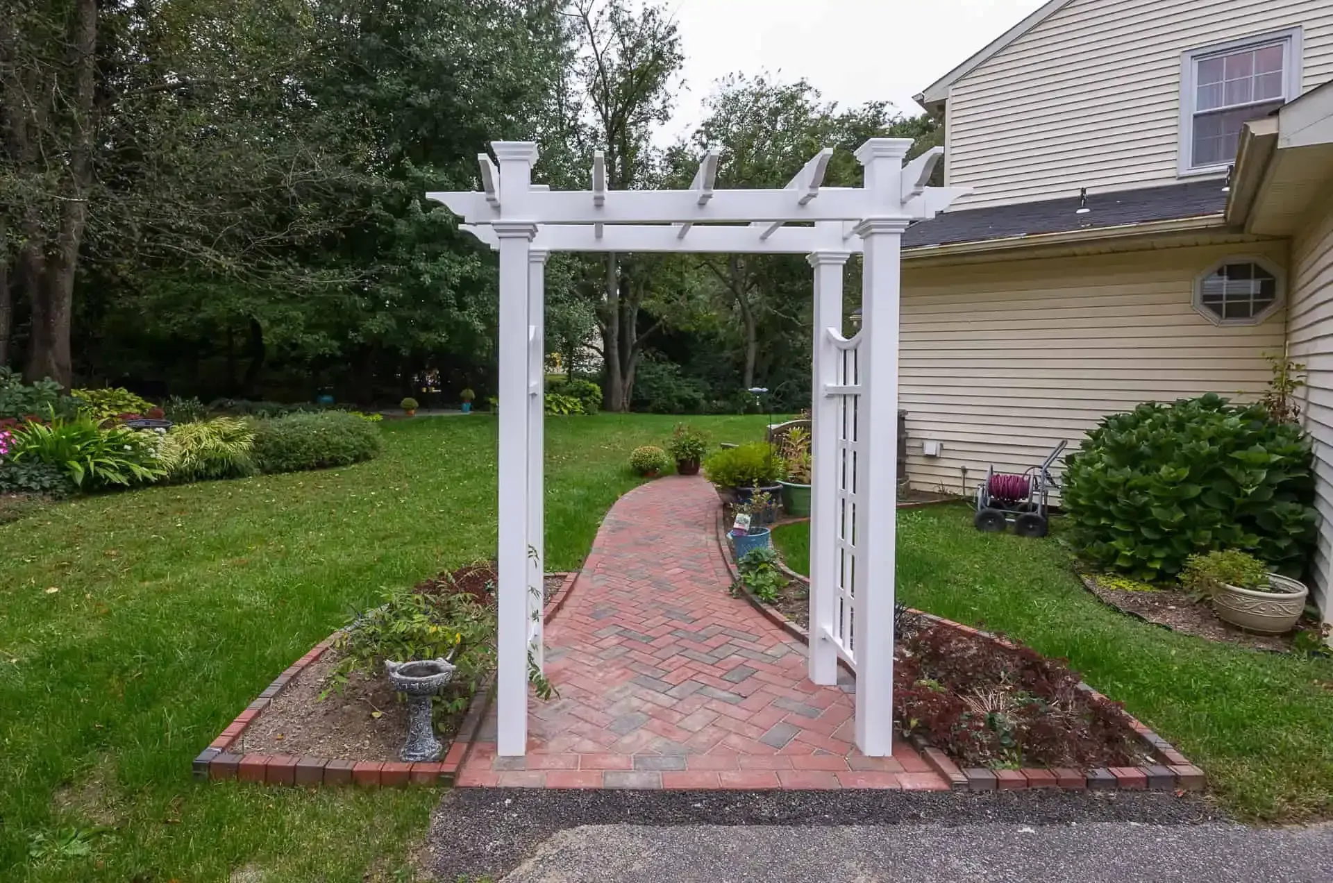 White arbor over a red brick garden path beside a beige house and green lawn