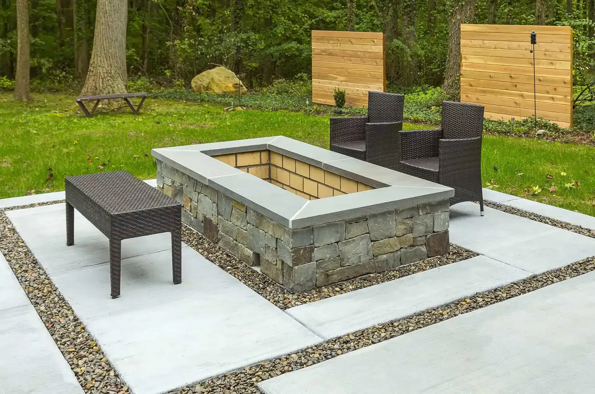 Stone fire pit seating area with black wicker chairs on a concrete patio in a wooded yard