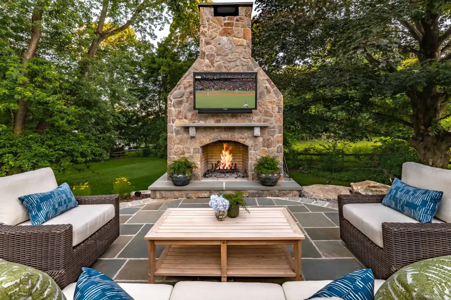 Outdoor patio with stone fireplace, cushioned chairs, and a wooden coffee table in a lush garden