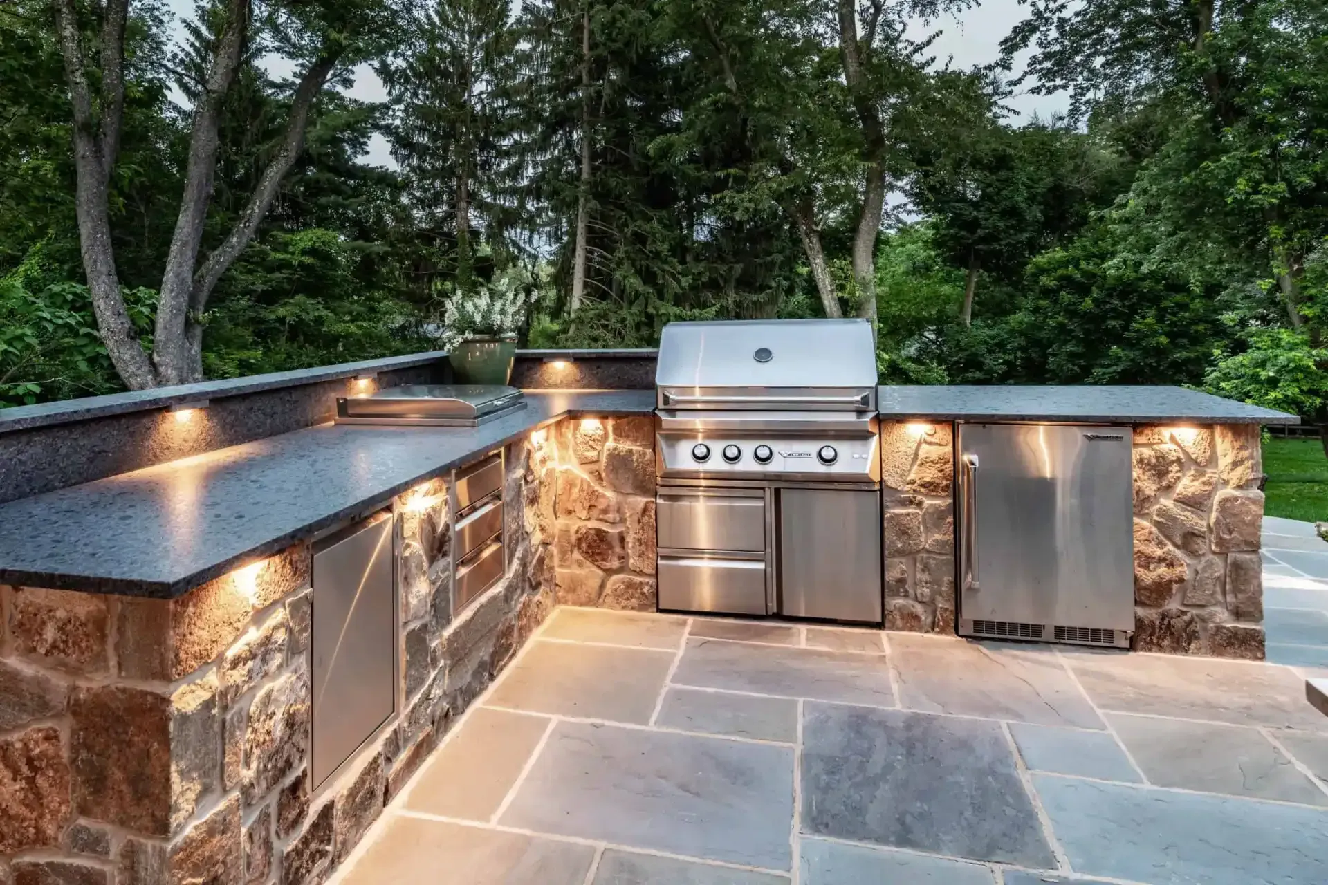 Outdoor stone patio kitchen with built-in grill, sink, and cabinets under trees at dusk