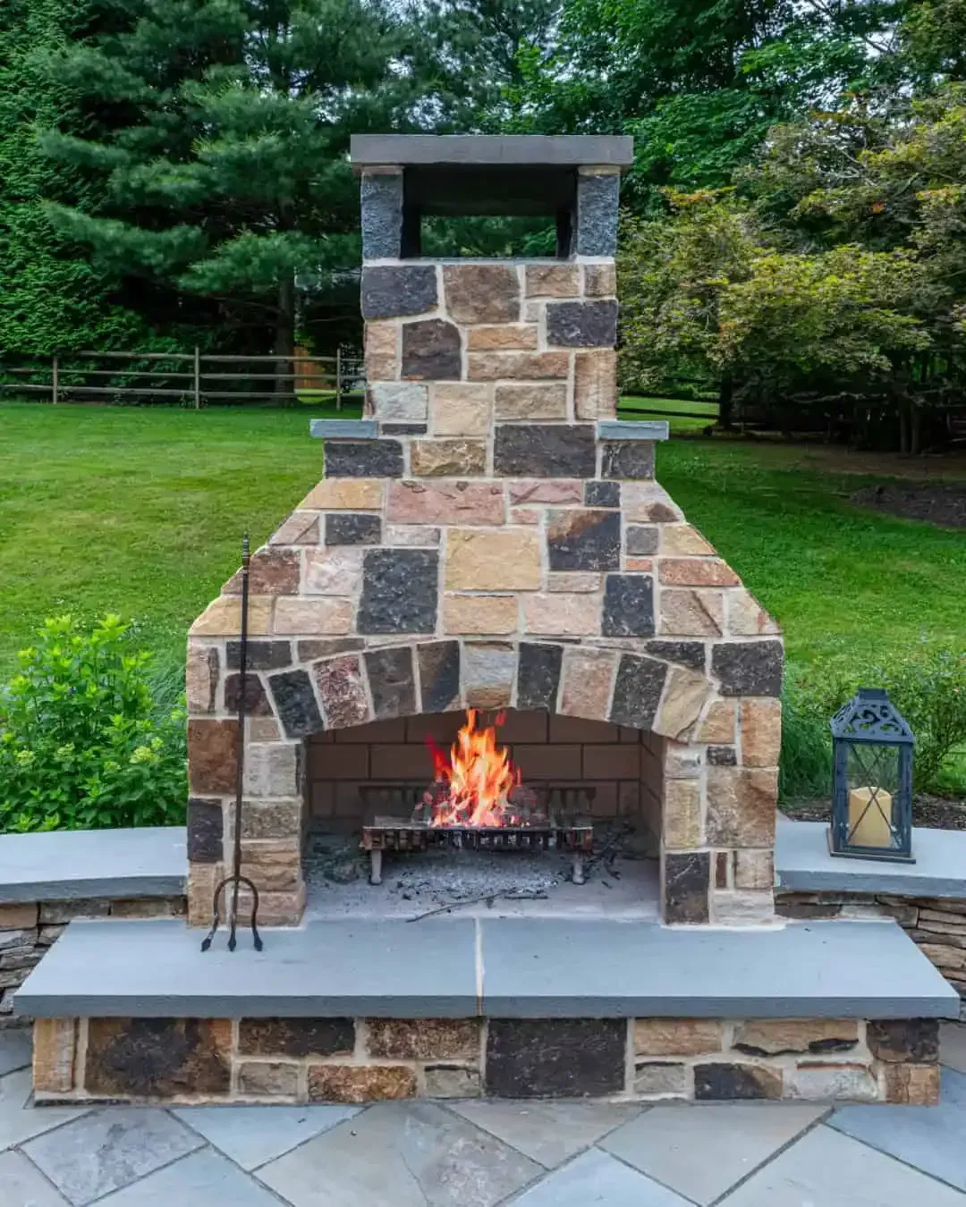 Stone outdoor fireplace with a lit fire, set in a grassy backyard with trees and a lantern nearby