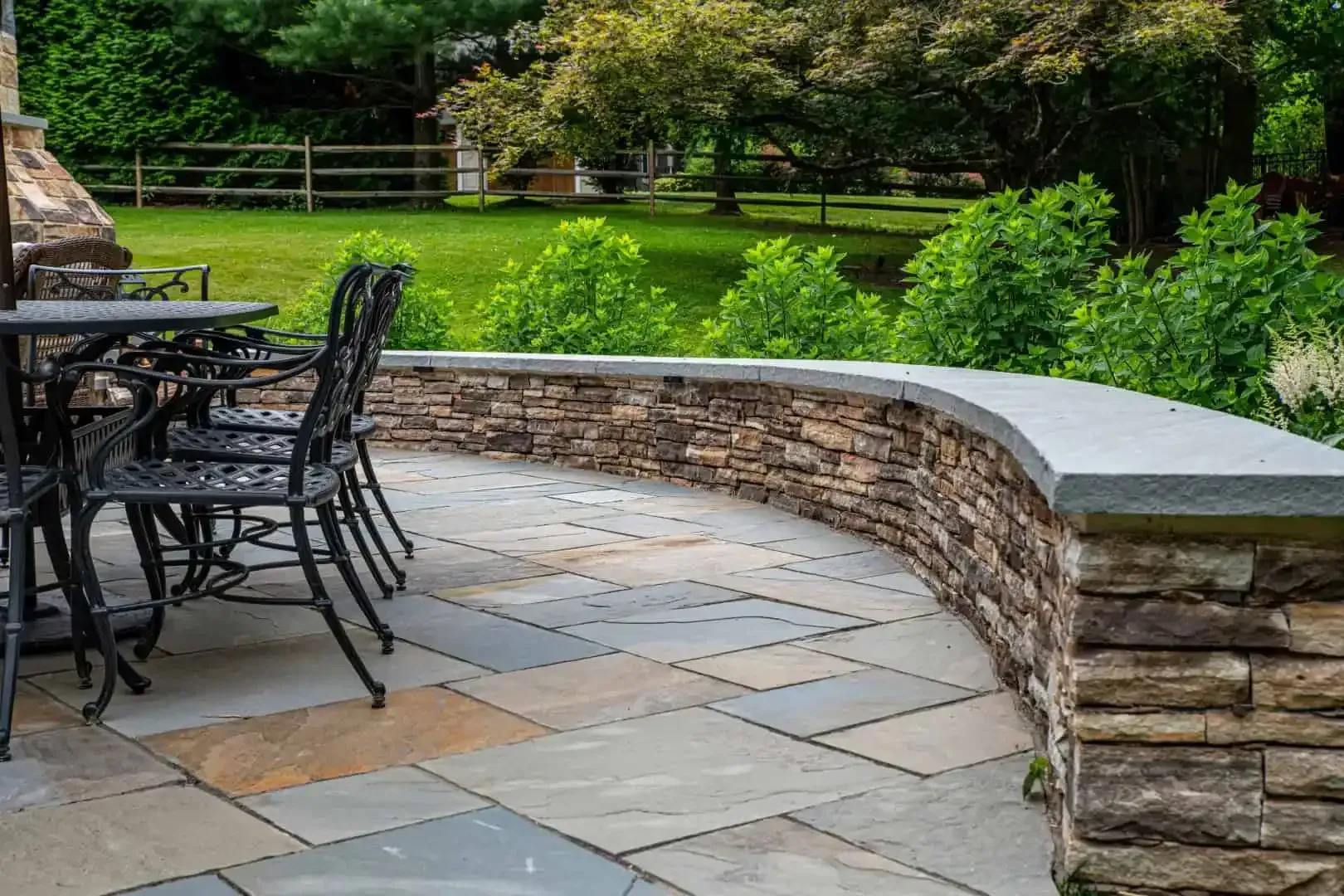 Stone patio with black metal chairs and a curved retaining wall beside a lush green garden