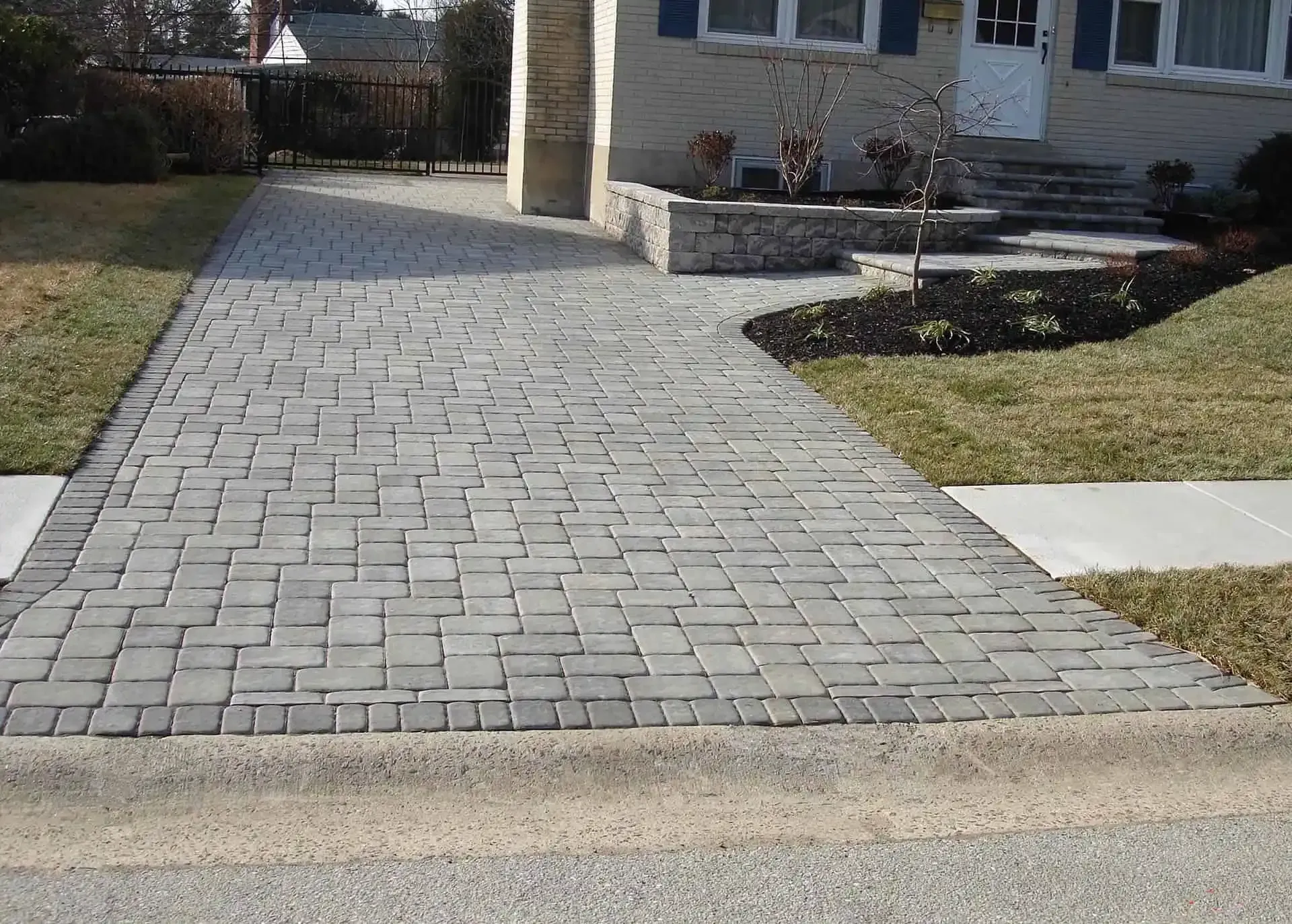Brick driveway leading to a house entrance, bordered by grass and a concrete sidewalk.