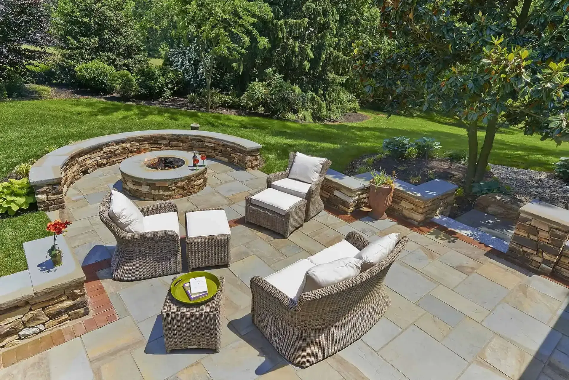 Stone patio with wicker seating, round fire pit, and lush green backyard garden trees