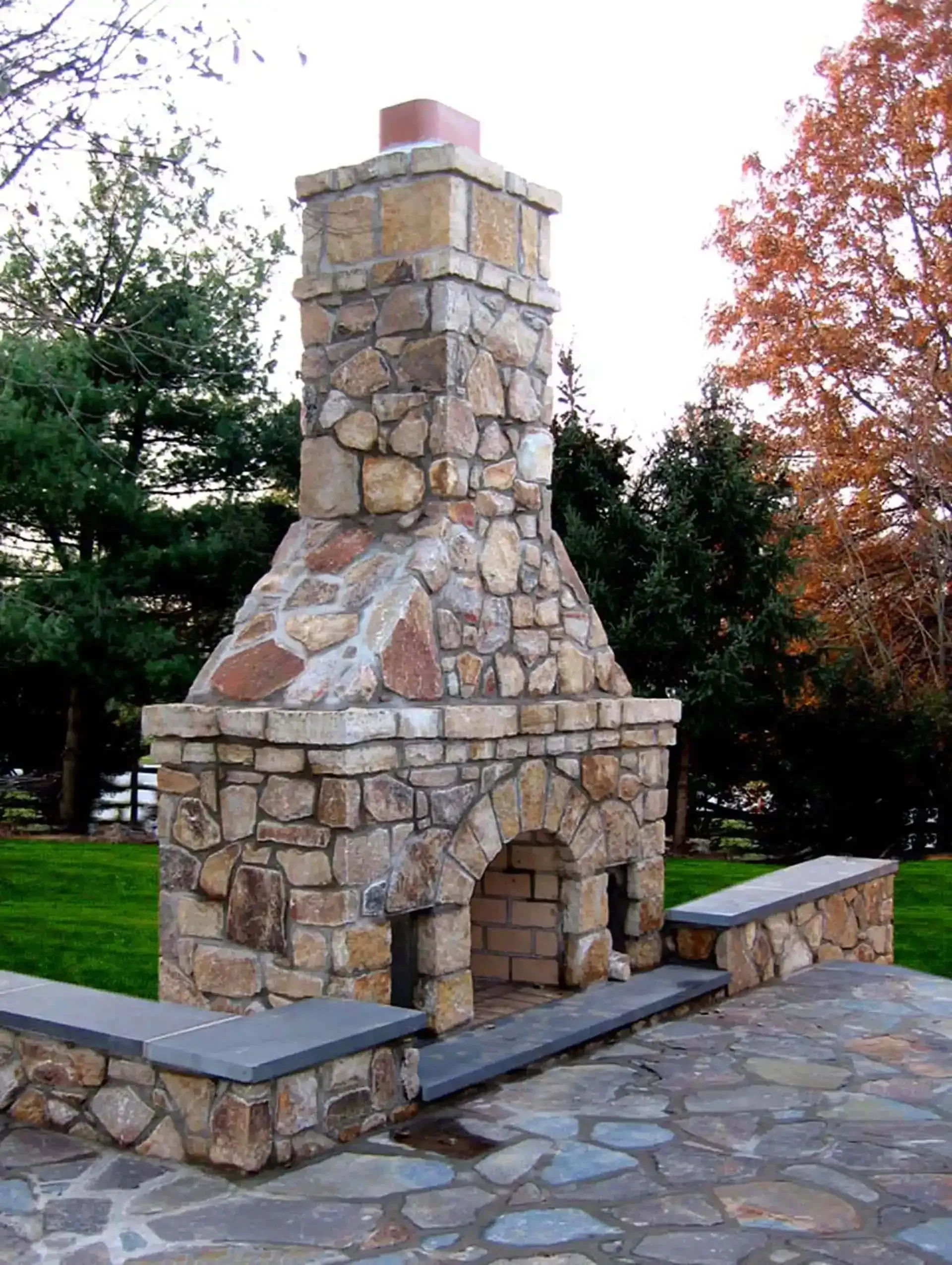 Stone outdoor fireplace with benches on a paved patio in a park setting