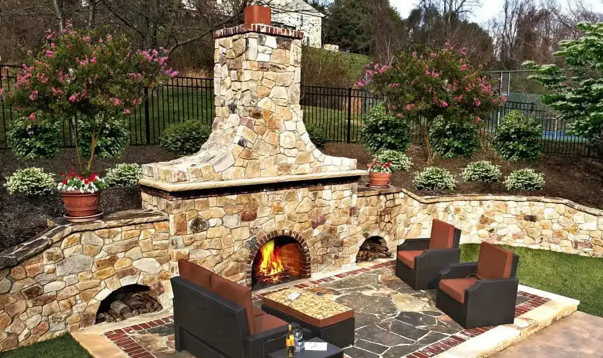 Stone outdoor fireplace with burning fire and brown patio chairs in a landscaped garden