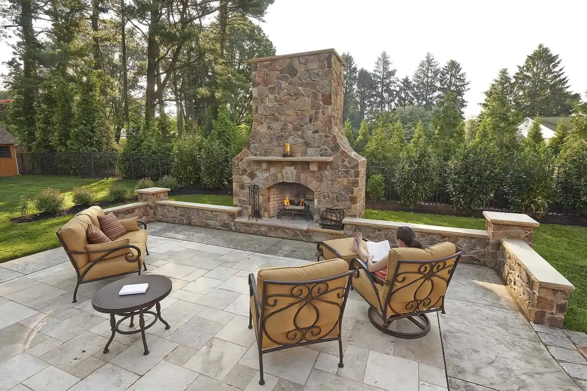 Stone patio with cushioned chairs around a rustic outdoor fireplace in a landscaped backyard