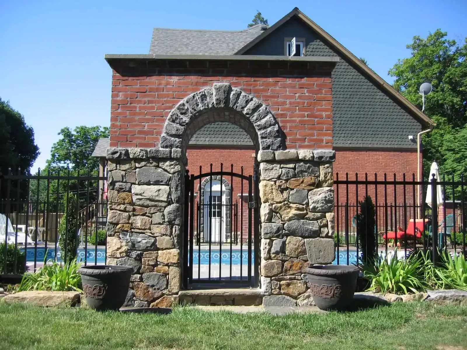 Stone garden gate with arched brick entry and wrought-iron fence beside a pool, with plants and houses behind