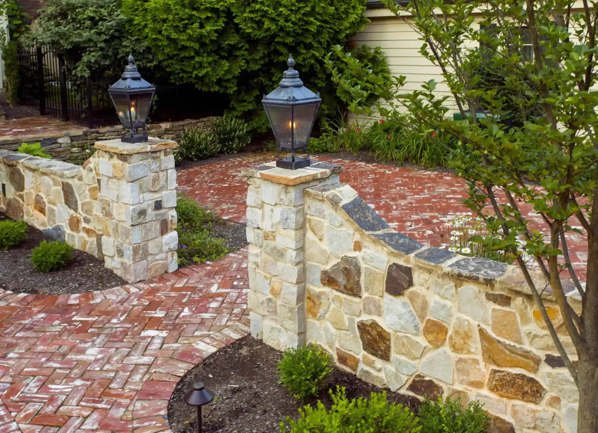 Stone garden wall with black lanterns and a red brick walkway among greenery