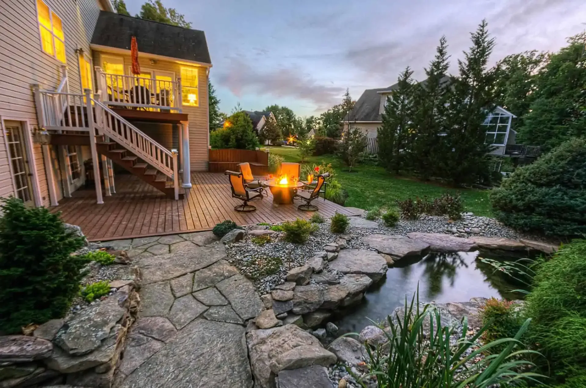Cozy backyard patio with stone path, fire pit, pond, and lit house deck at dusk