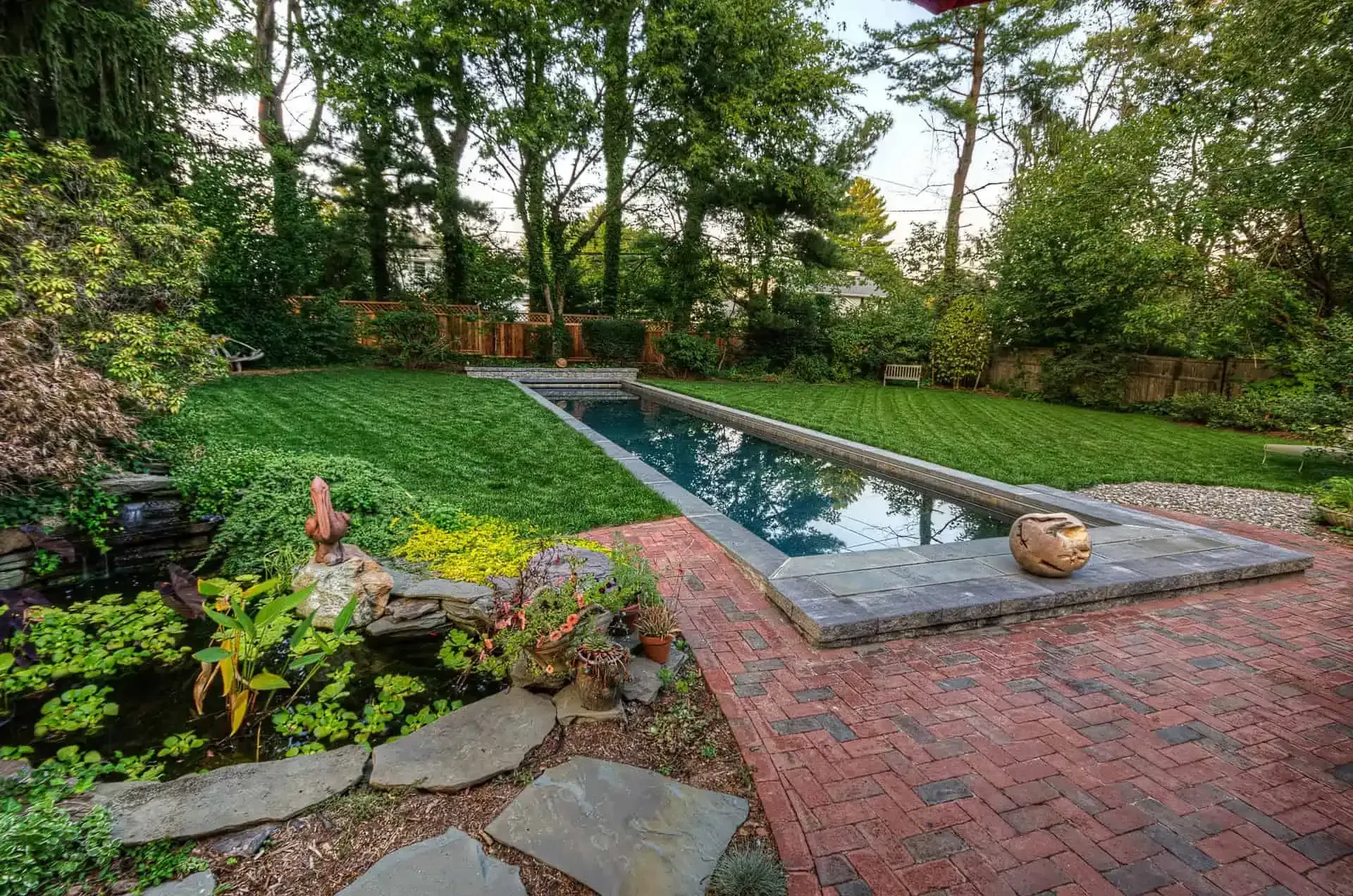 Backyard with a rectangular reflecting pool, red brick patio, stone edging, and lush greenery