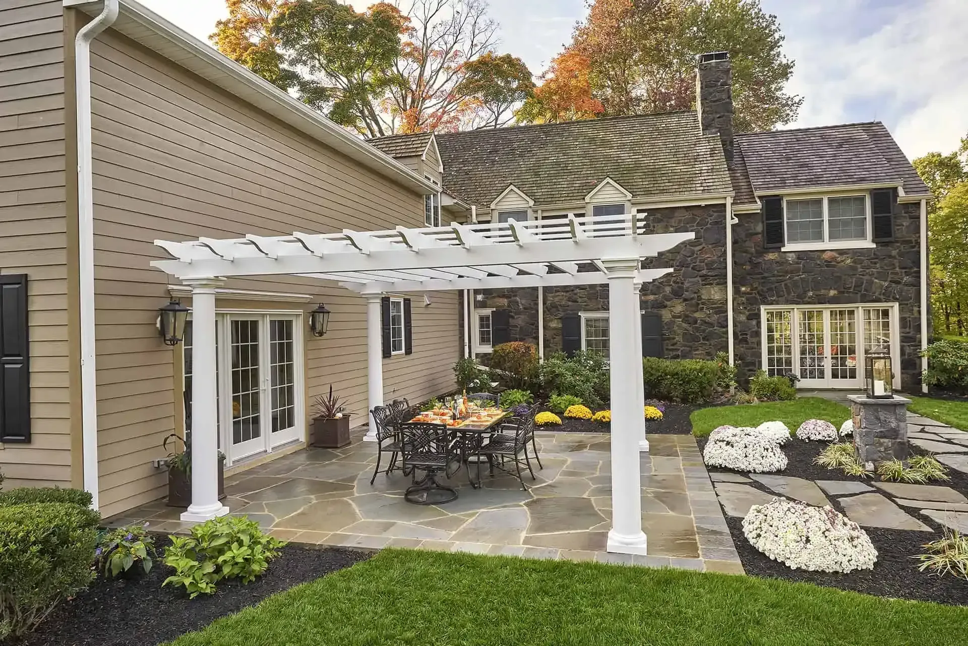 Patio with white pergola, outdoor dining set, stone paving, and landscaped garden beside a house