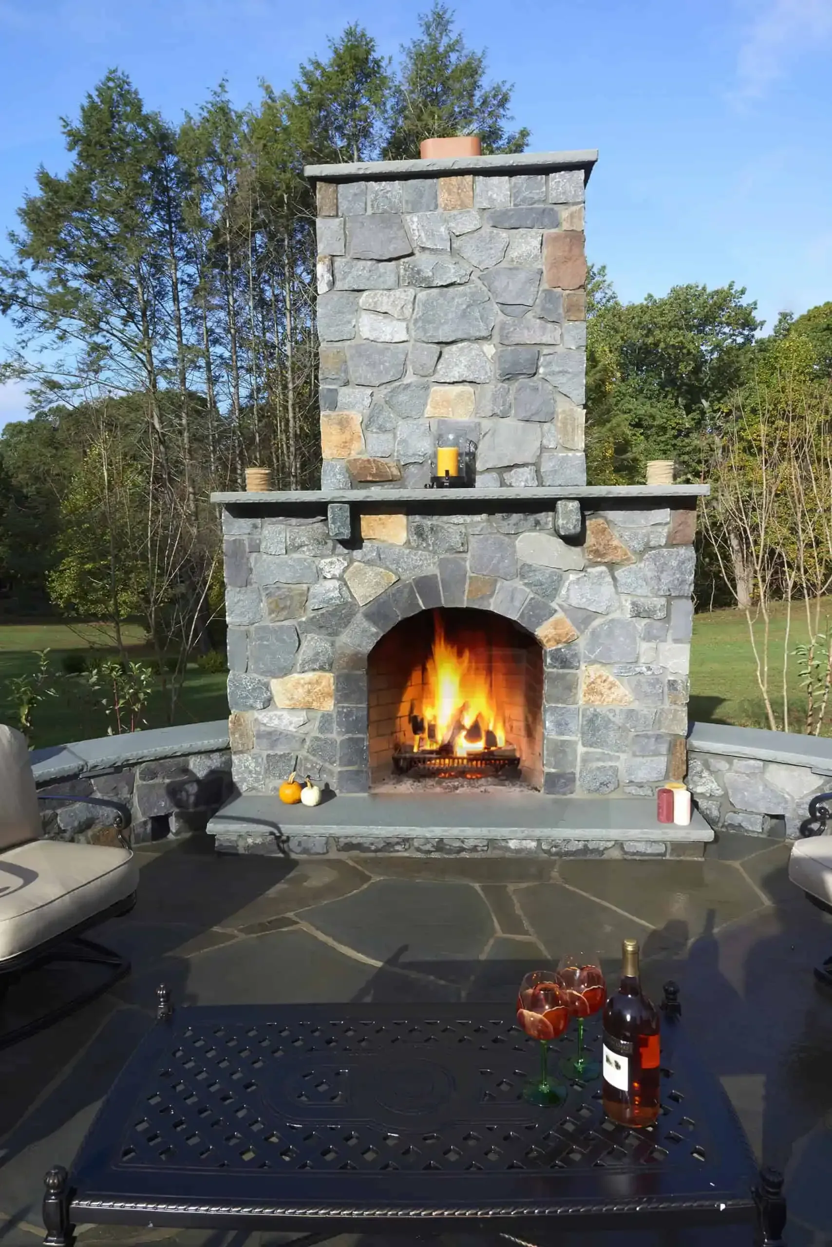 Stone outdoor fireplace with a lit fire on a patio, surrounded by seating and trees in the background