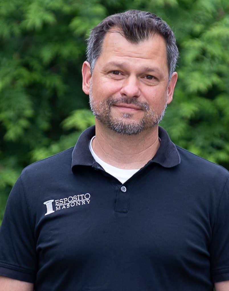 A man in a black polo shirt standing outdoors in front of green foliage, smiling at the camera