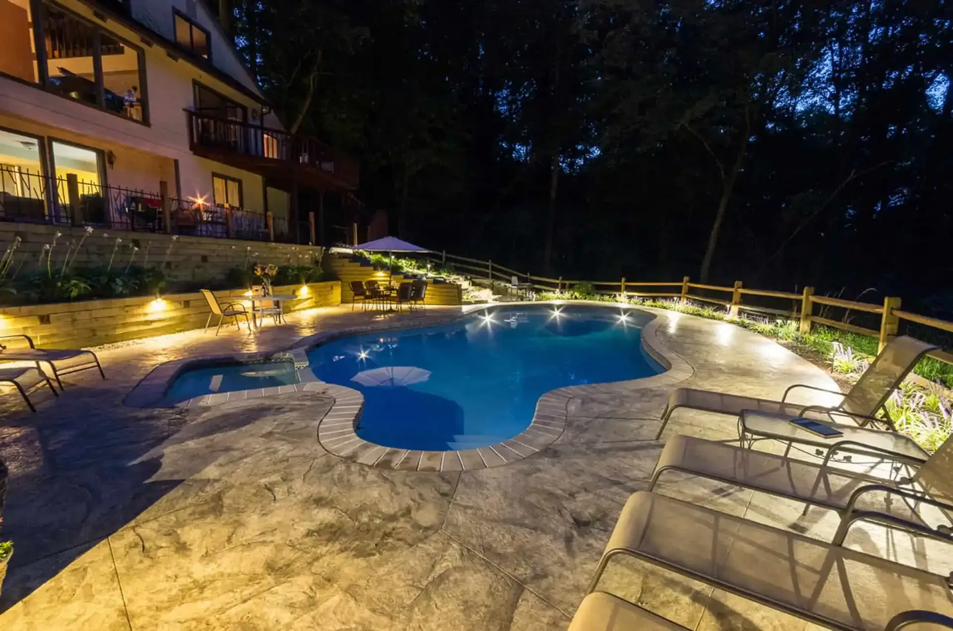 Nighttime resort pool with blue water, stone deck, and lit lounge chairs beside a building and trees