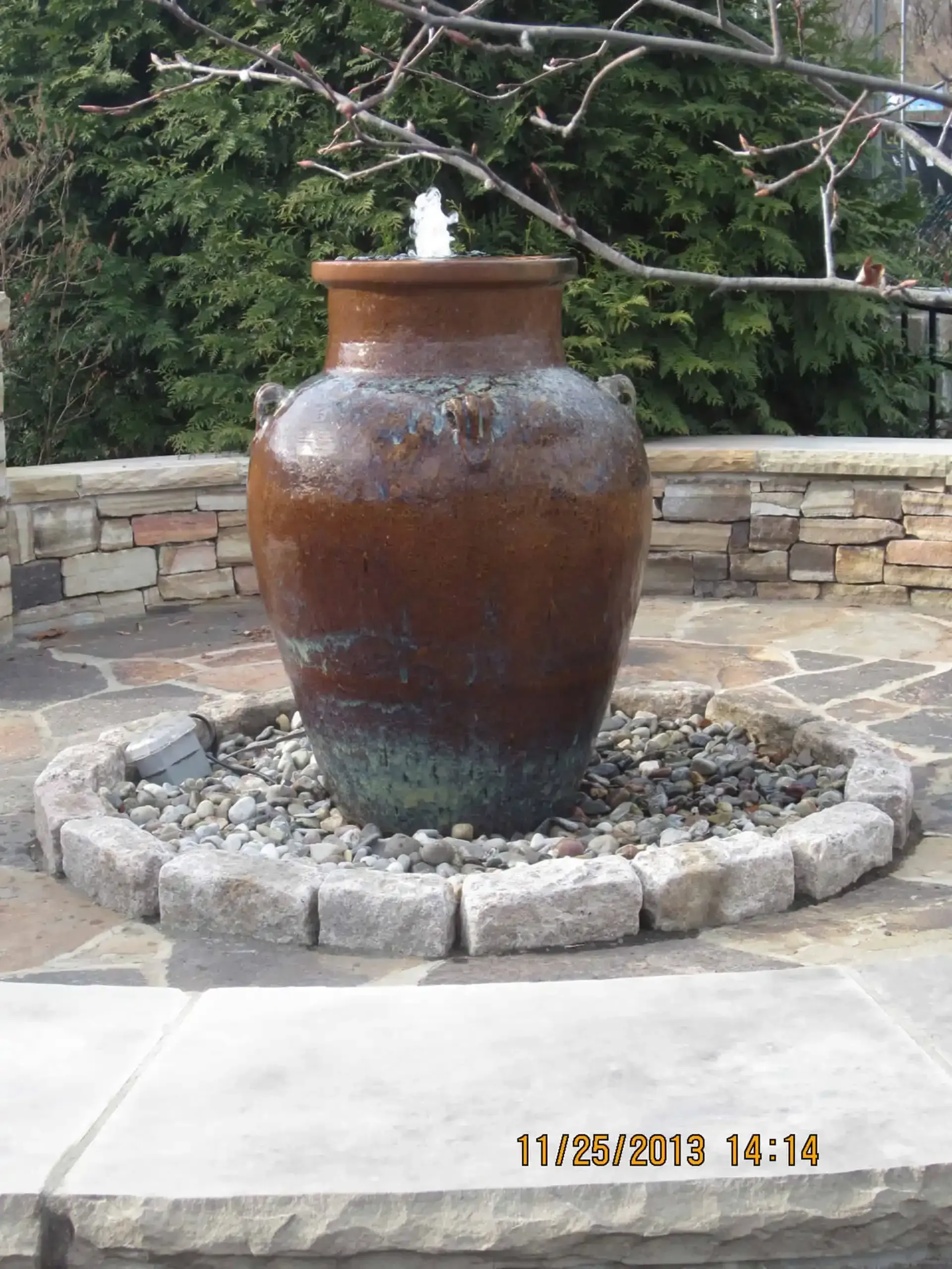 Large brown ceramic urn fountain in a stone courtyard with water flowing over the top.
