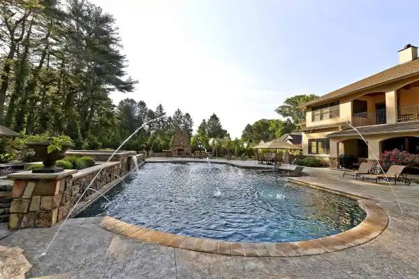Backyard pool with stone deck and two-story house, surrounded by trees and lounge chairs
