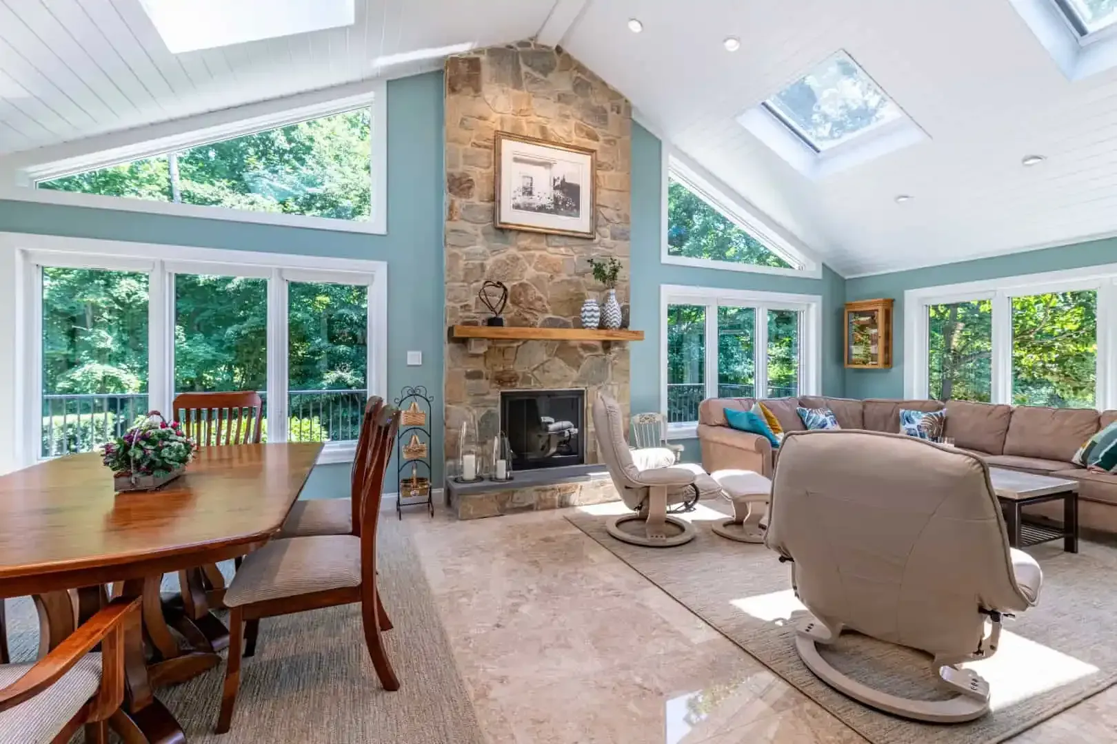 Bright living room with vaulted ceilings, stone fireplace, skylights, and large windows overlooking trees