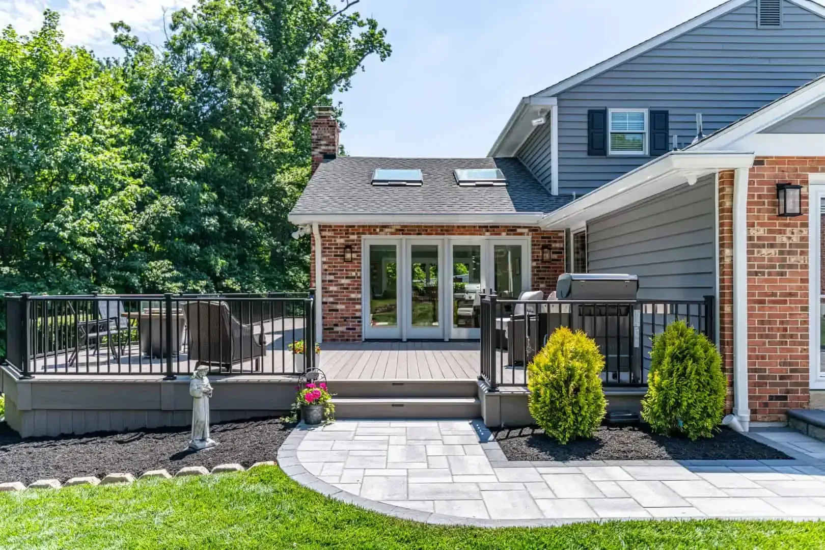 Backyard patio with gray pavers, black railing, potted flowers, and a gray house with French doors