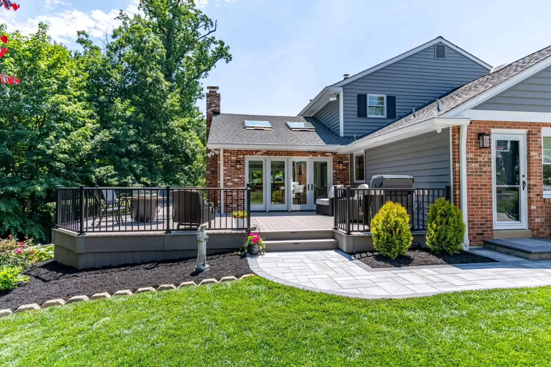 Backyard patio with gray siding, stone walkway, deck railing, and green lawn beside a house