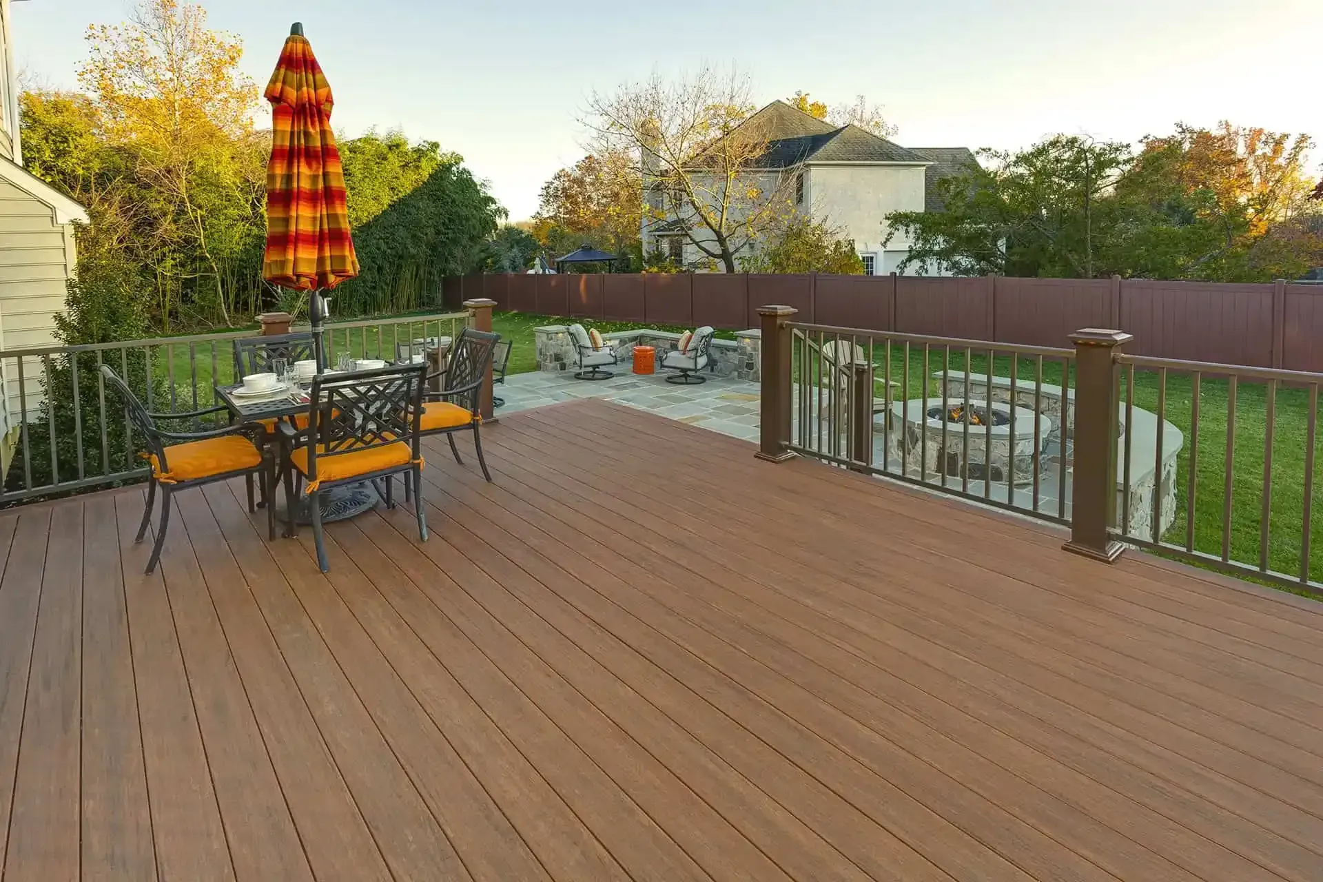 Spacious wooden deck with patio table, striped umbrella, and glass railing overlooking a yard at sunset