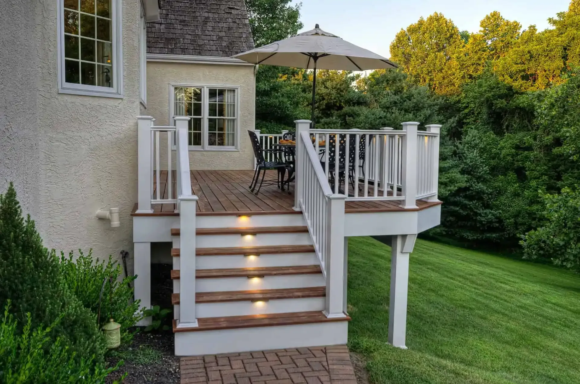 Backyard deck with lit stairs, white railing, patio table, and green lawn beside a house