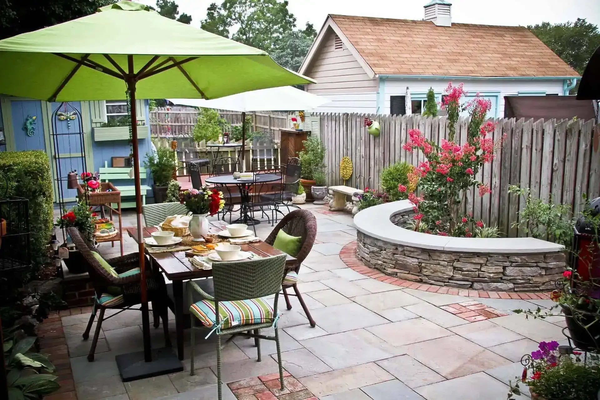 Patio dining area with green umbrella, table setting, potted flowers, and stone fire pit in a backyard garden
