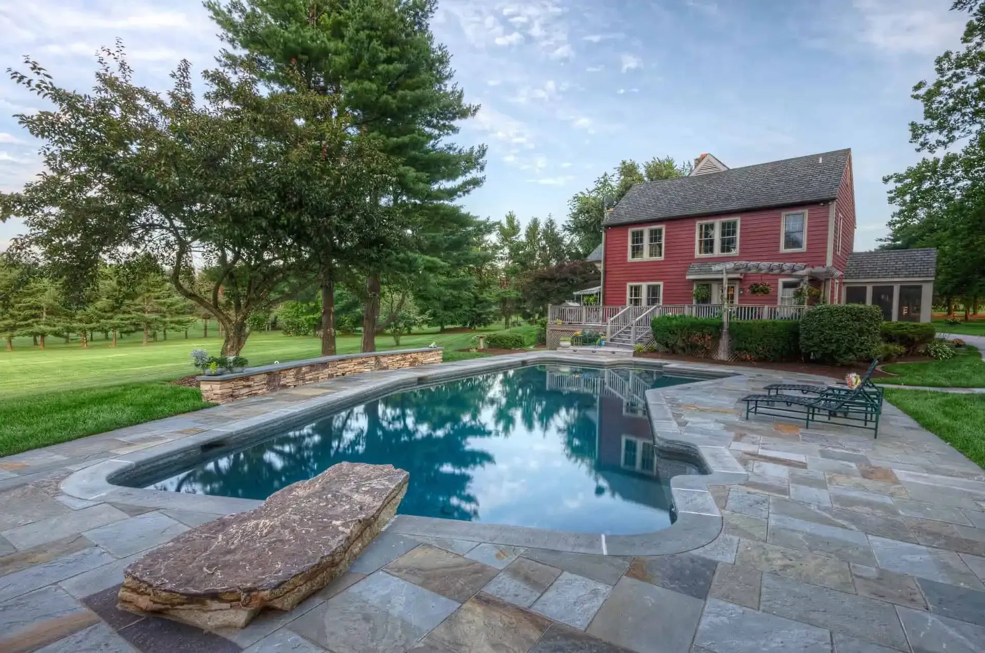 Backyard pool with stone patio beside a red two-story house and green lawn